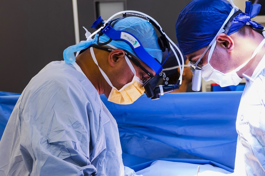 Two doctors in blue medical scrubs.  The doctor on the left is holding surgical tools and operating on a patient.