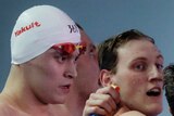 A male swimmer wearing a swimming cap walks past a group of swimmers celebrating winning a relay event.
