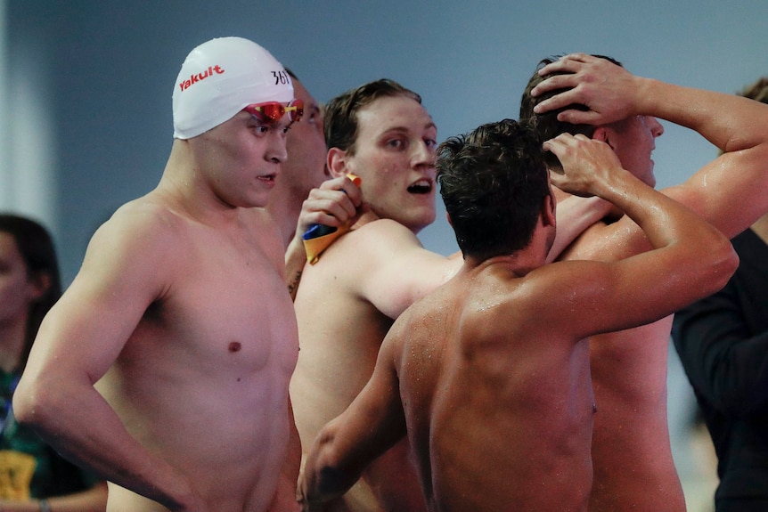 A male swimmer wearing a swimming cap walks past a group of swimmers celebrating winning a relay event.