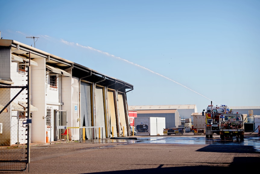 A firefighting truck sprays water at a fire-damaged warehouse, on a sunny day.