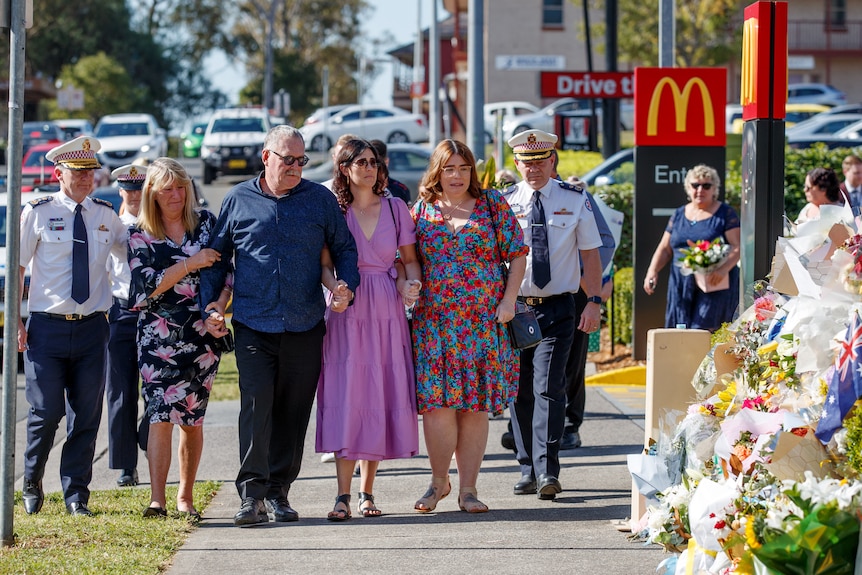 Four family members walk arm in arm with emergency service workers in front of floral tribute 
