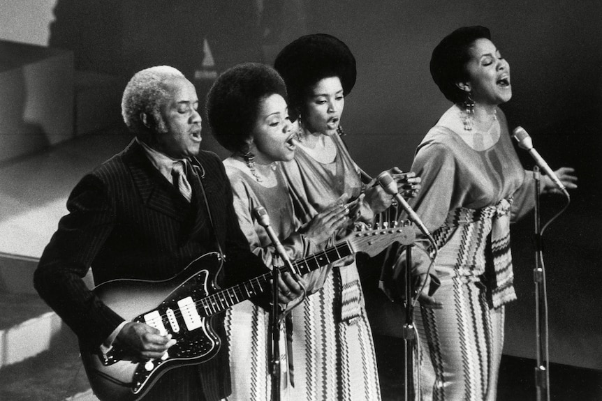 A black and white photo of a Black man holding a guitar and singinging standing next to three Black women singing in the 70s