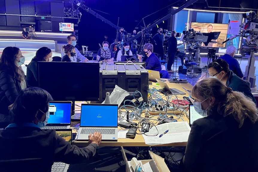 Desk with eight people working on laptops in a TV studio with hosts and cameras in the background.