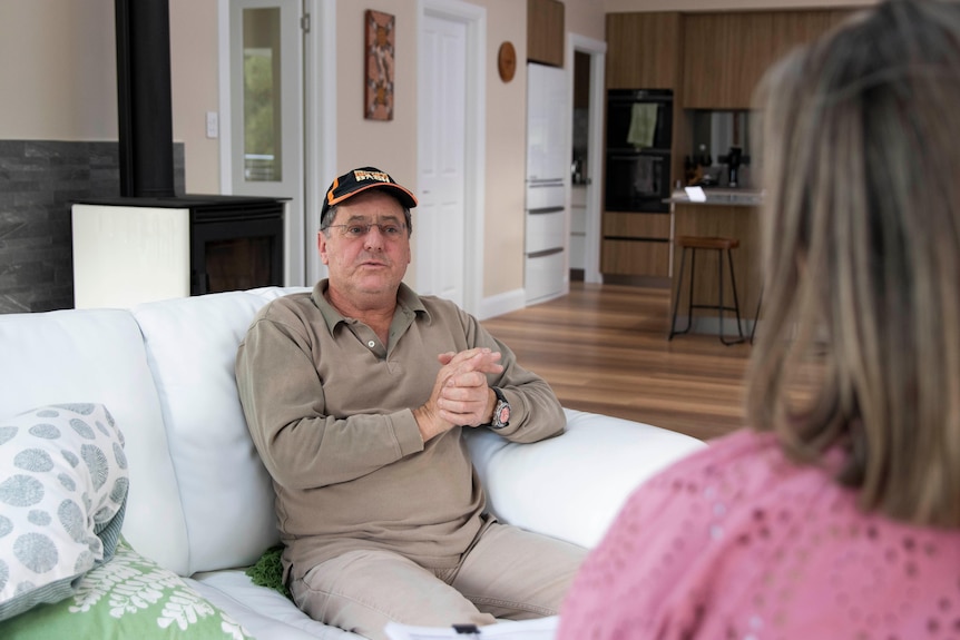 A man sits on a couch talking to a reporter with her back to the camera.