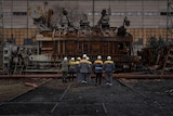 a group of power plant workers in hard hats and protective gear walk together towards wreckage in a Ukrainian power plant