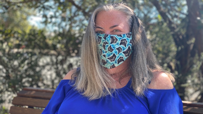 A woman with long ash-blonde hair sits on a bench under a tree, wearing a mask.