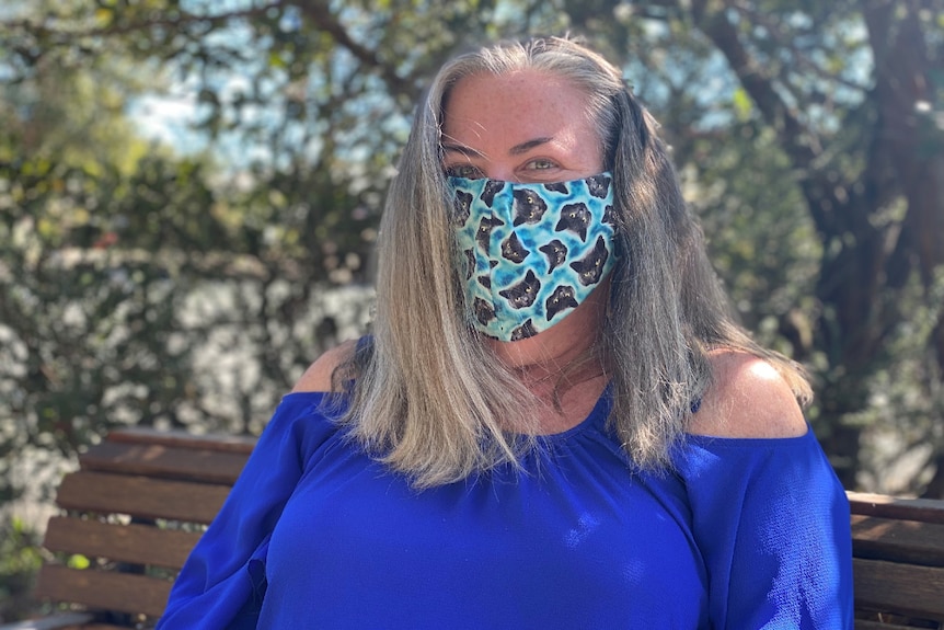 Woman sits on a chair outside under a tree wearing a mask