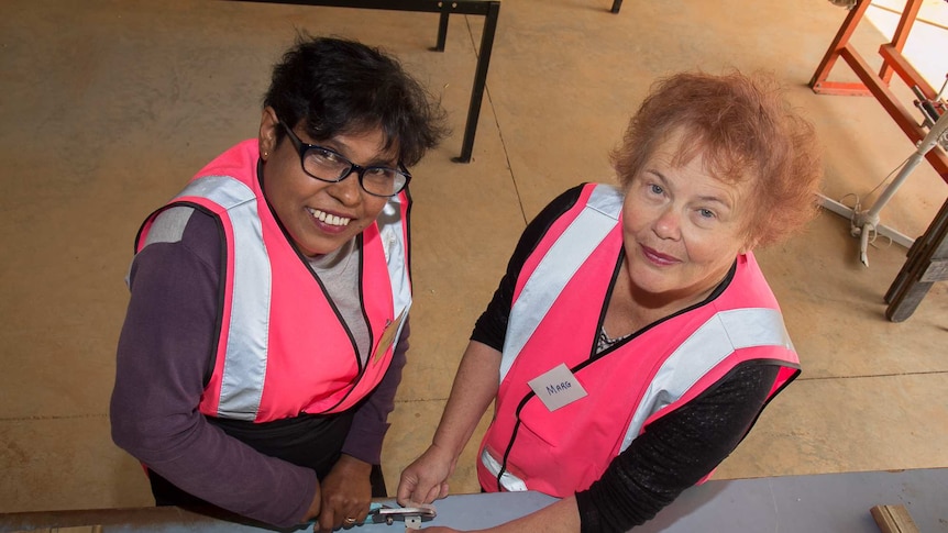 Two women in bright pink high visibility vests using tools on a workbench with wood around
