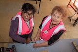 Two women in bright pink high visibility vests using tools on a workbench with wood around
