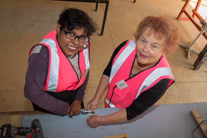 Two women in bright pink high visibility vests using tools on a workbench with wood around