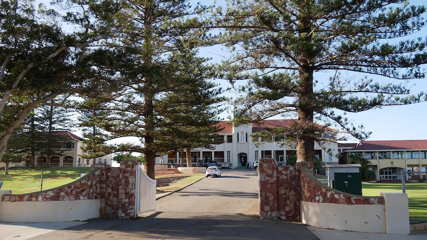 The driveway leading up to a large two-storey Catholic school with large trees in the foreground.