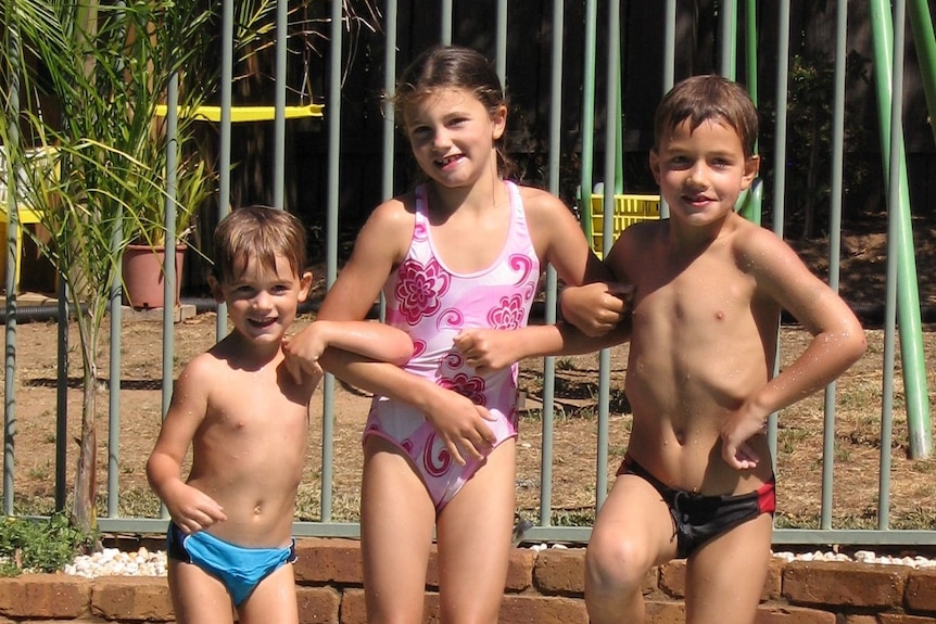 Trois enfants souriants, une fille, deux garçons, dans des baigneurs posant pour une photo près d'une piscine.