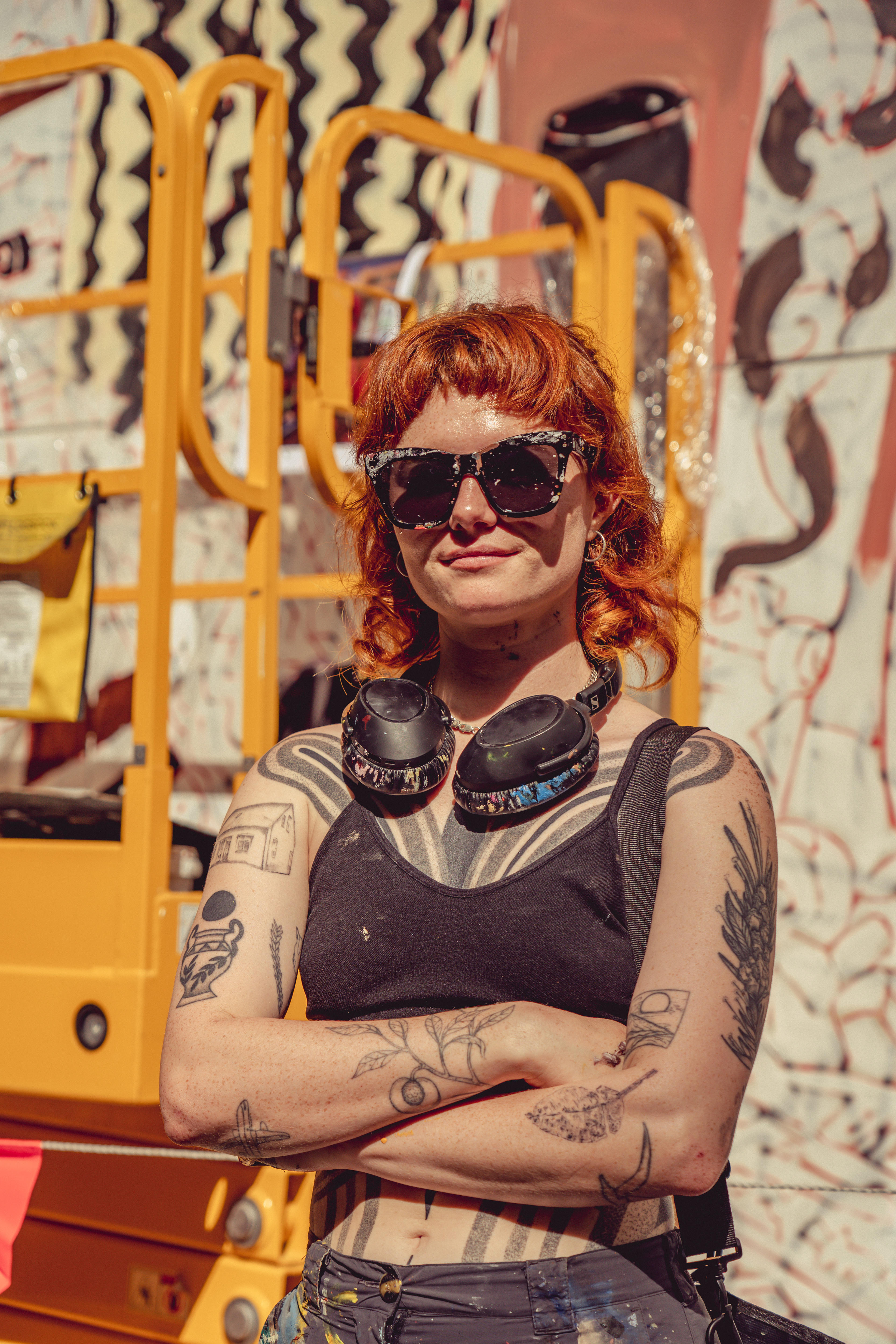 Photo of the artist looking at the camera, with arms closed - they are in their late 20s with fair skin, red hair and sunglasses