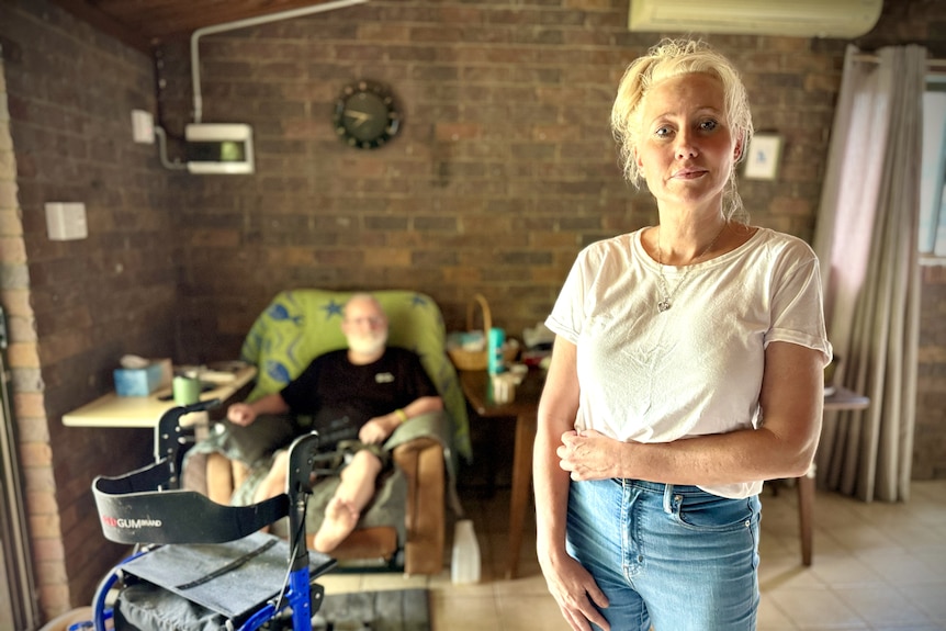 A woman stands staring at the camera with her father in a recliner behind her and a walker in front of him.
