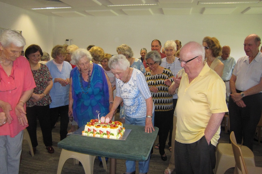 A woman blowing out a 100th birthday cake with dozens of people surrounding her