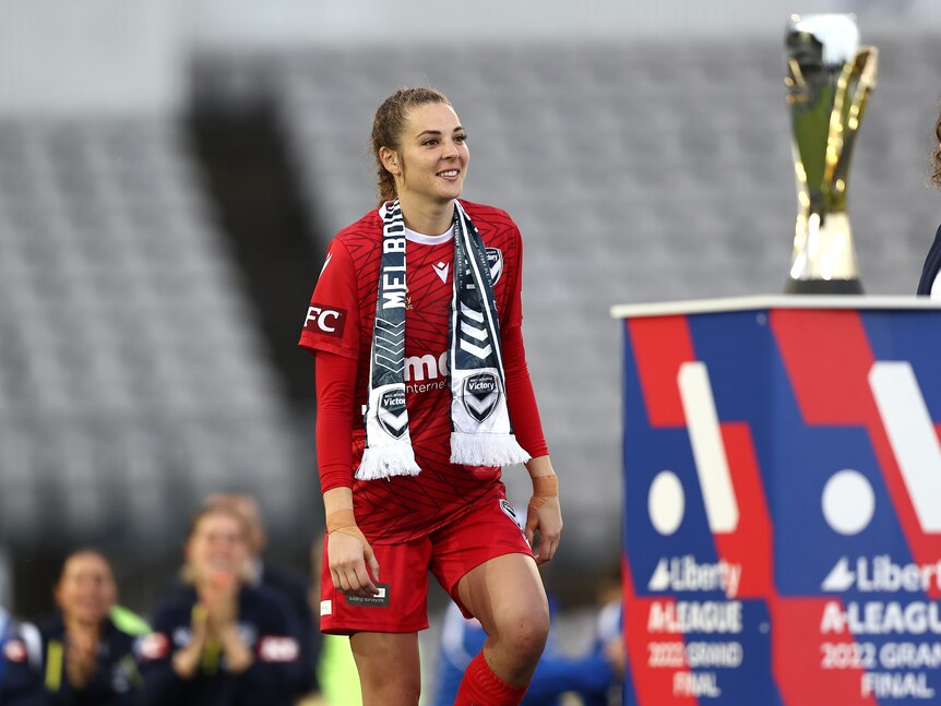 A soccer player wearing red and a blue scarf walks on stage to receive a medal