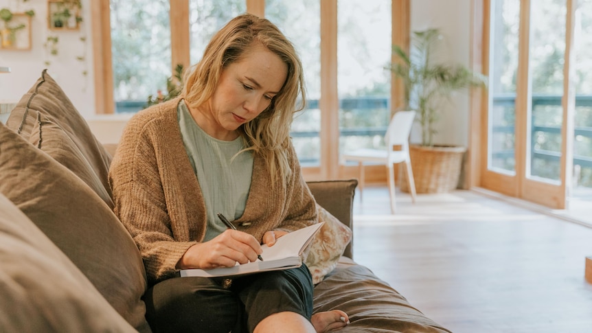 A woman sits on a couch, writing in a journal.