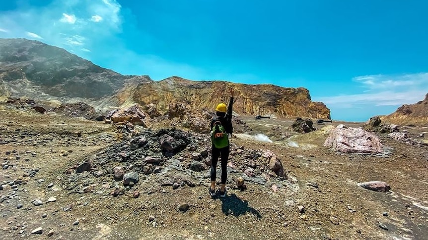 A woman in a hard hat has her back to the camera and stands on Whakaari. 