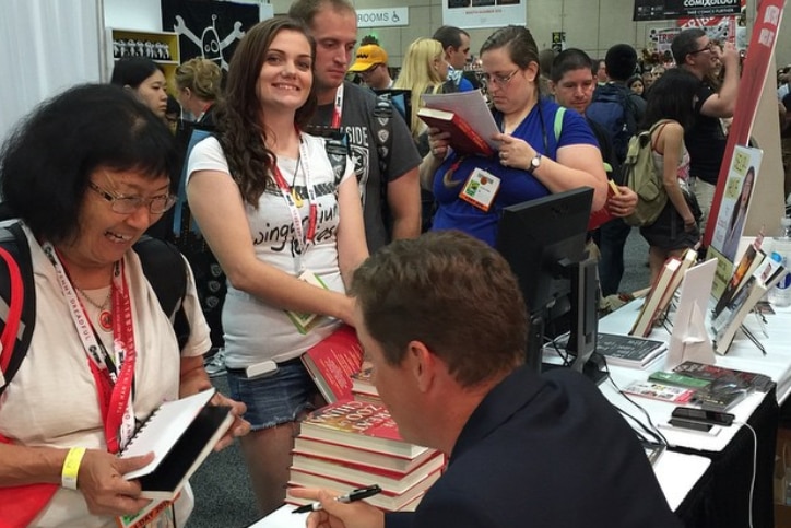 People line up in a crowded hall waiting to see a seated man who is signing books