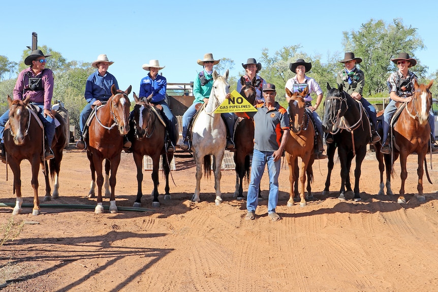 A group of people on horses smiling, with a man in front of them standing on red dirt holding a 'No gas pipelines' sign.