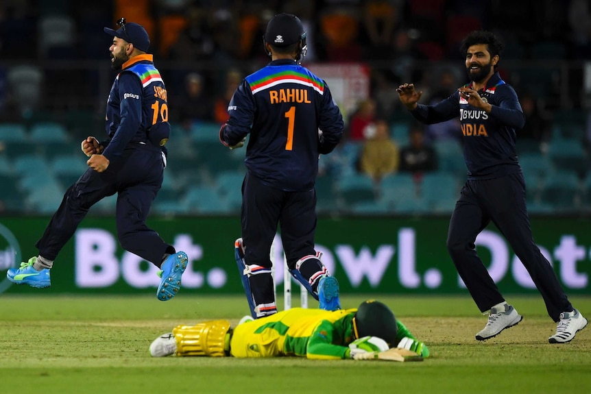 Three Indian male cricketers celebrate as a dismissed Australian batter lies on the ground.