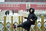 A woman stands in the snow with one leg up sin front of a Chinese building