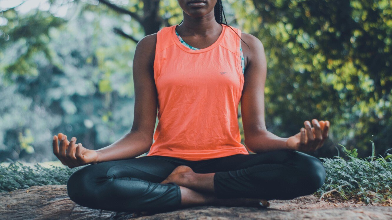 A woman wearing a red singlet sits down cross legged, her hands resting on knees, in a leafy park.