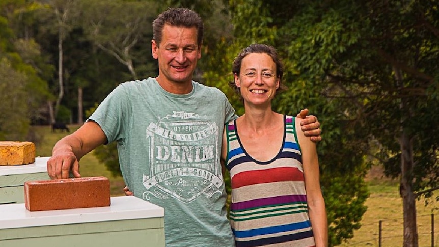 A middle-aged man stands with his arm around a woman's shoulders beside some beehives.