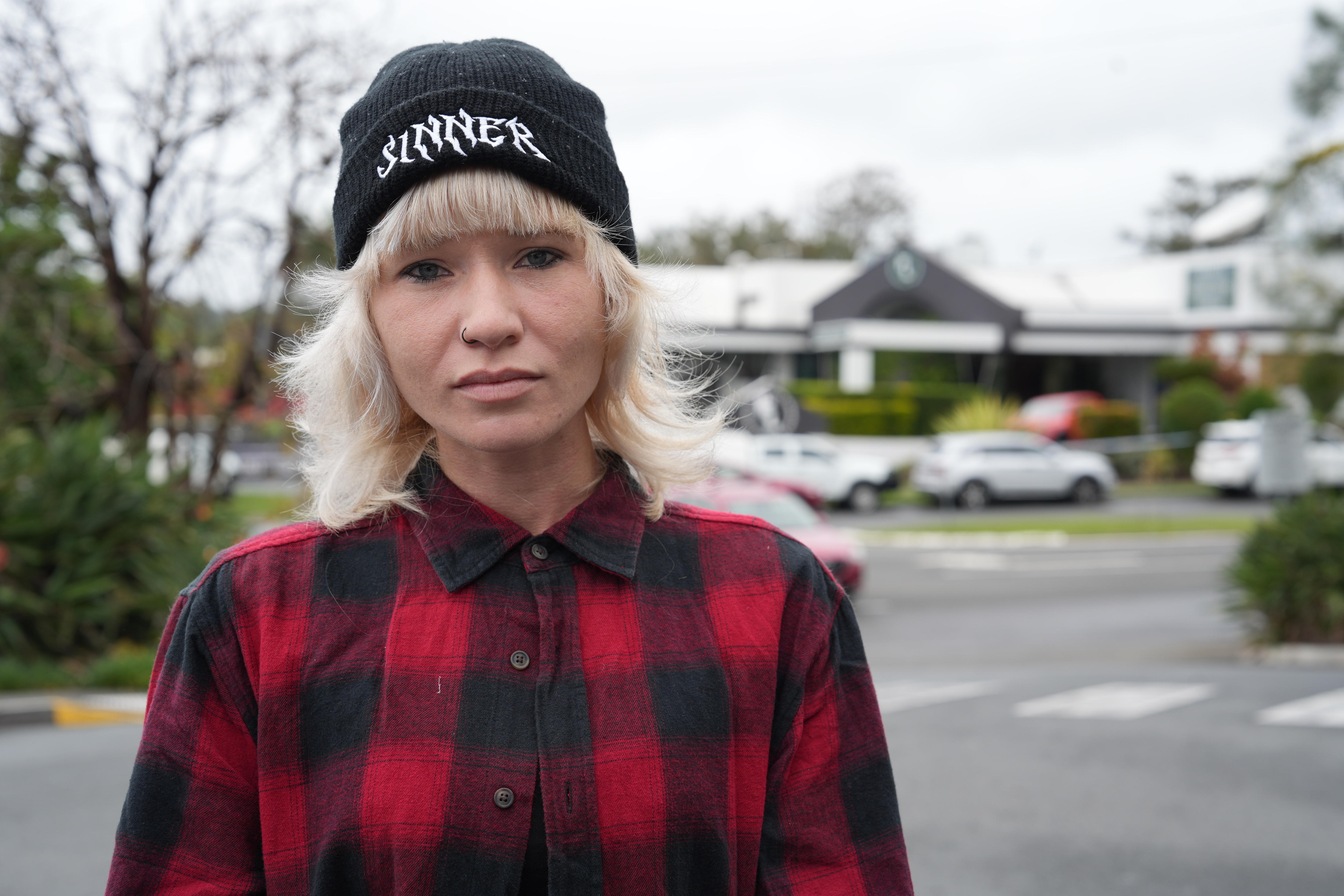 A woman with a blonde fringe in black beanie and red flannel shirt standing in car park.