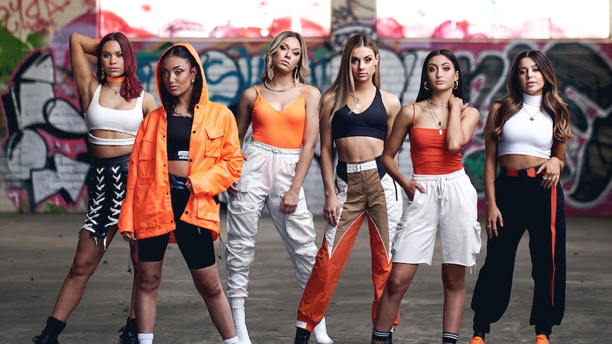 A group of female singers clad in orange and black pose while standing in a warehouse.