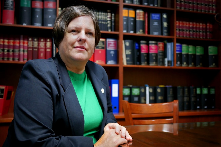 A woman in a green shirt and blazer with a large bookshelf behind her.