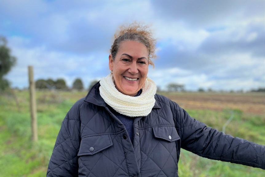 A smiling older with her hair tied back, but messy, wears white scarf, blue puffer jacket, stands in front of a fence.