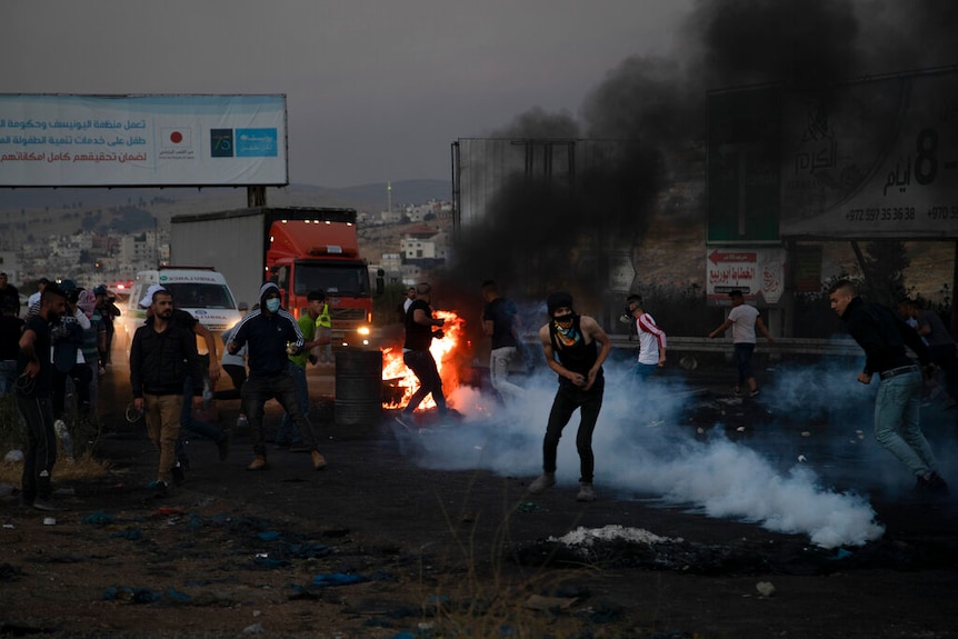 Mobs clash with military forces at a Middle Eastern check point. Fire and smoke can be seen on the road.