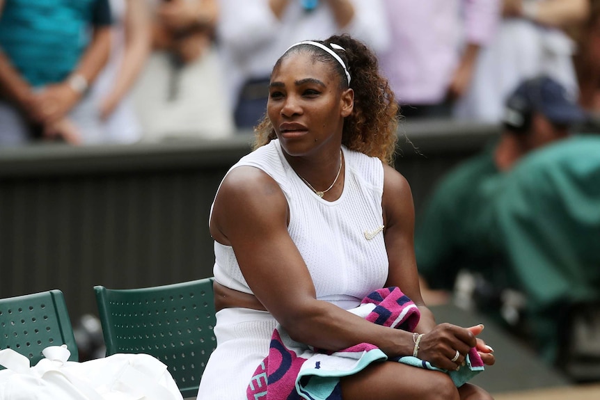 Serena Williams looks across while sitting on a chair beside a tennis court.