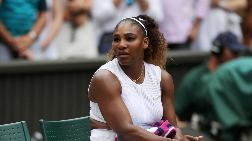 Serena Williams looks across while sitting on a chair beside a tennis court.