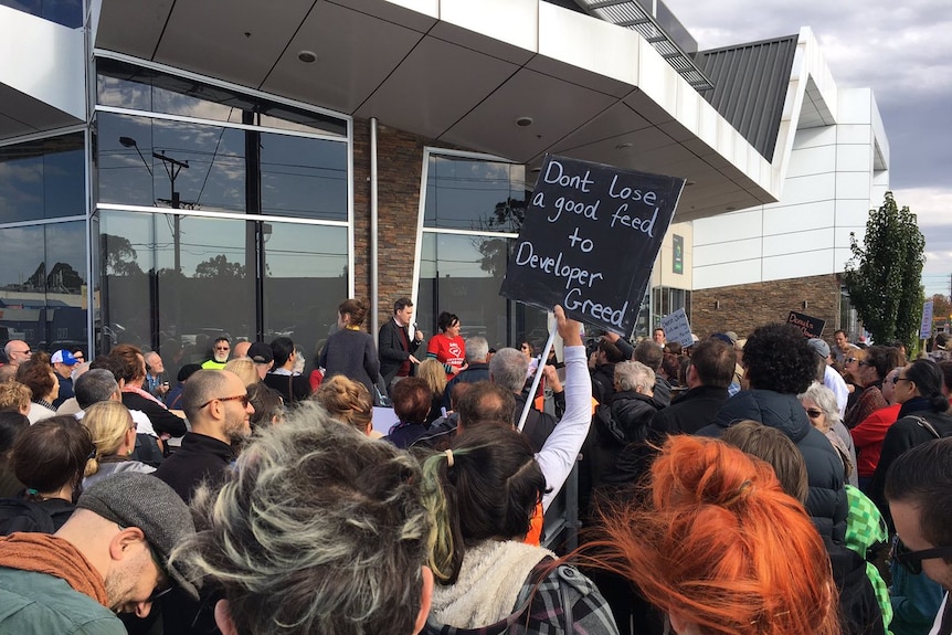 A large crowd looks on at a speaker at the front, as a man holds a sign that says 'Don't lose a good feed to developer greed'.