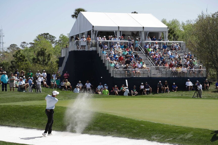 A golfer splashes out of the bunker, as the crowd watches from a greenside grandstand.