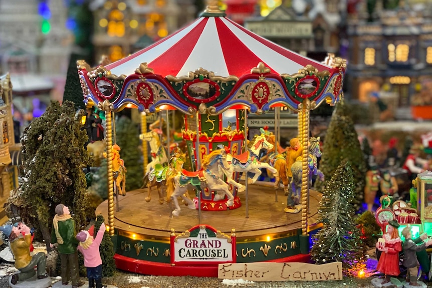 Miniature carousel with Christmas theme decorated village in background, lit up with lights.