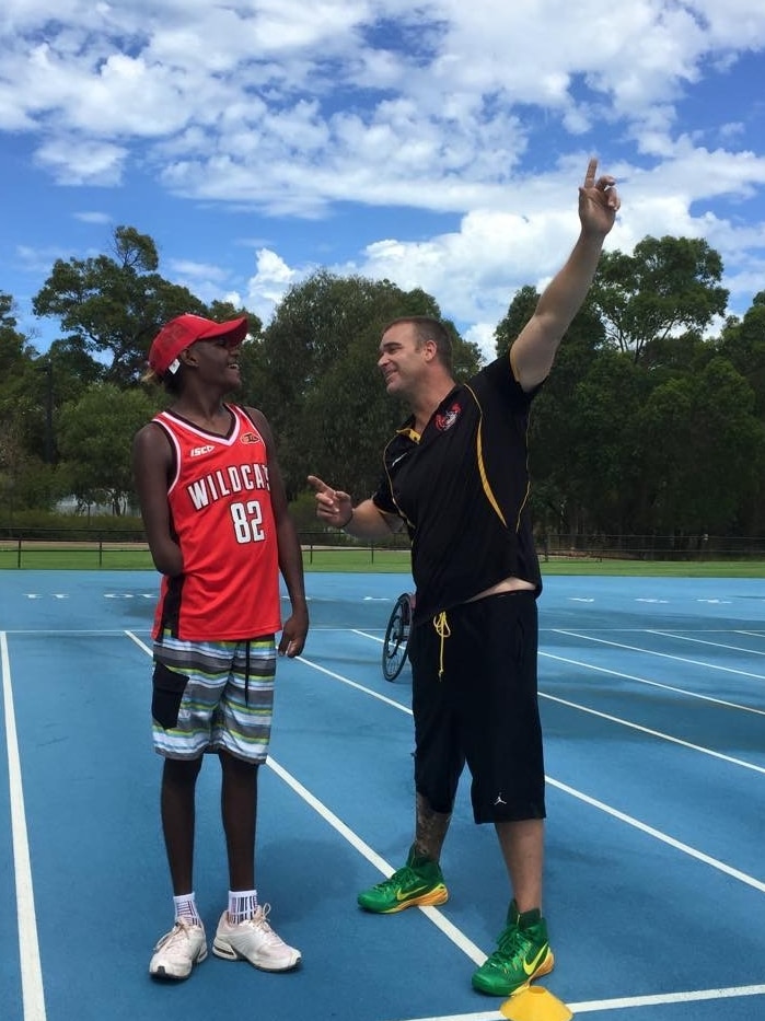 A coach points ahead for a younger man as the two stand at the start of a running track.