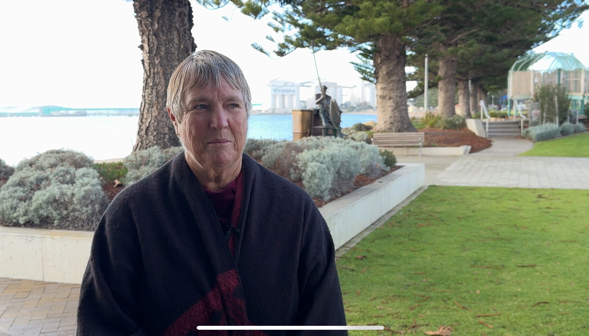 A woman wearing a navy-coloured jumper and scarf stands near the ocean.