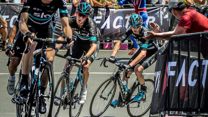 Team Sky riders make their way up a hill past the crowd, who are leaning over barriers and applauding the riders.