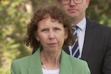 A woman wearing a green blazer stands with a serious expression, behind her is a man in a suit