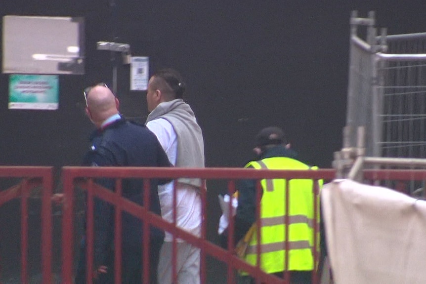 A man is escorted by police through the bakck of a building 