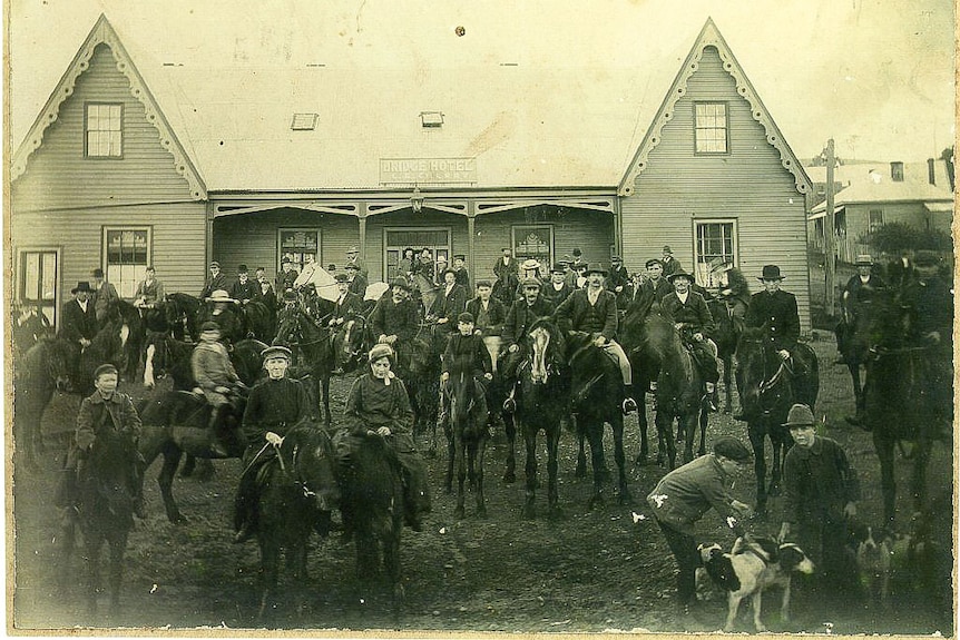 A crowd of men, women and children on horseback readied for a hunt, hotel building behind.
