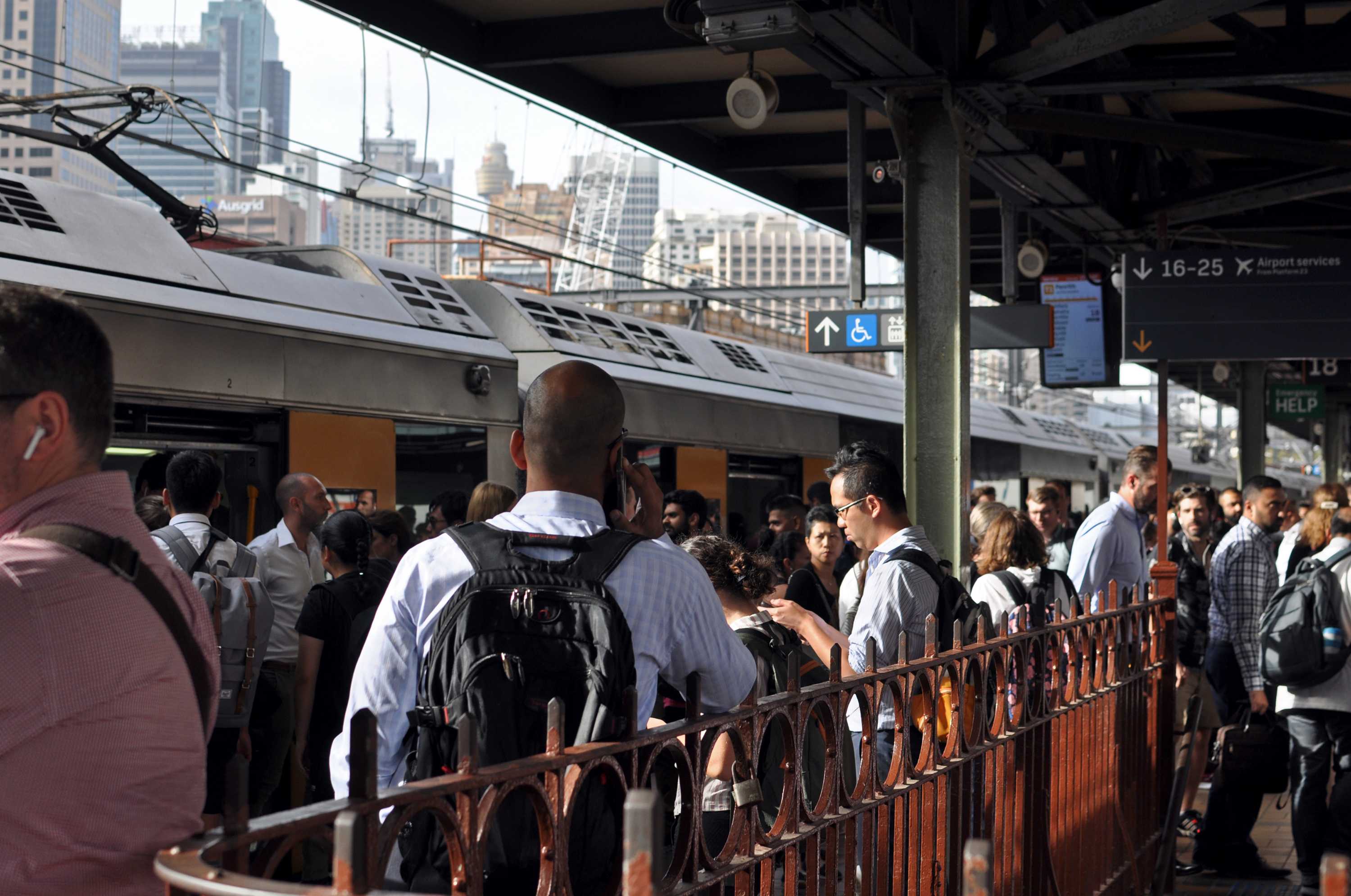 A crowded outdoor train station with people getting off and on of trains. Others talk and look at phones.