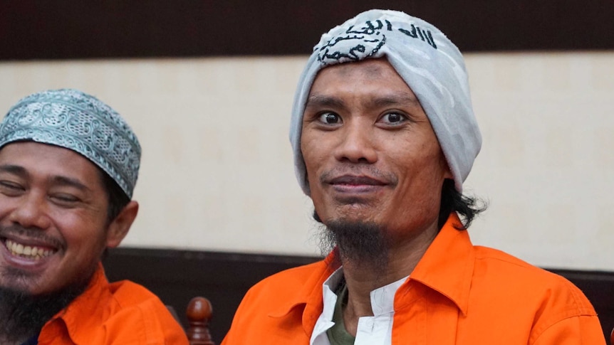 Juhanda sits in orange prison clothes smiling, as co-accused laughs next to him