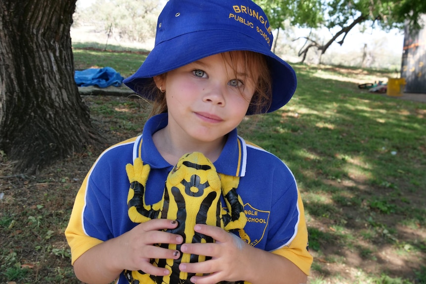Small child dressed in blue and yellow school uniform holds yellow and black toy frog.