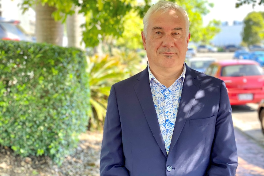 Man in a suit stands on a street with trees in background not looking happy