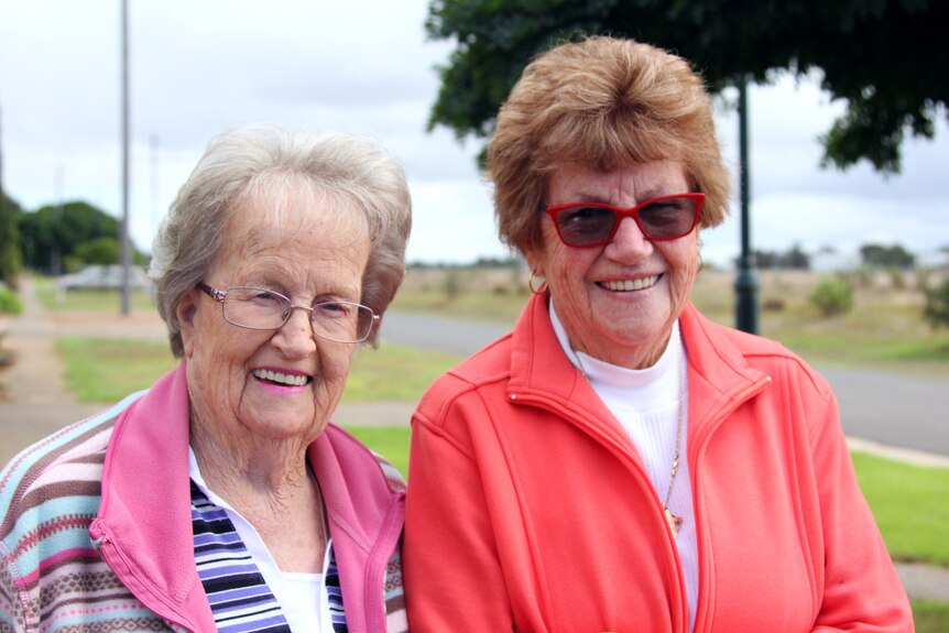 Two women in their 80s standing on a suburban street with a footpath on one side and paddocks on the other