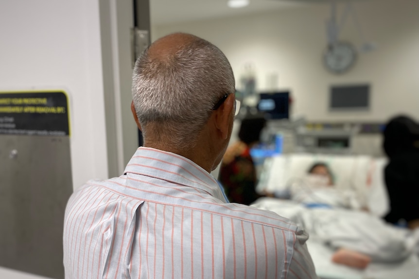 Fadel, wearing a business shirt with orange stripes, looks on into a hospital room where his daughter is being treated 